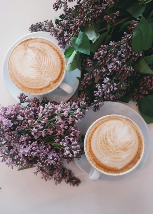 Artistic latte art surrounded by blooming lilac flowers creates a cozy, aesthetic vibe.